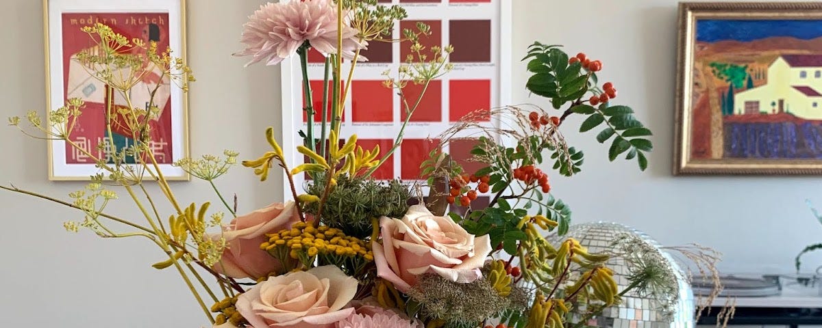 A large sunny bouquet of flowers, including pink roses and other pink and red flowers. The bouquet is on a table surrounded by small disco balls, and paintings are on the wall behind them.