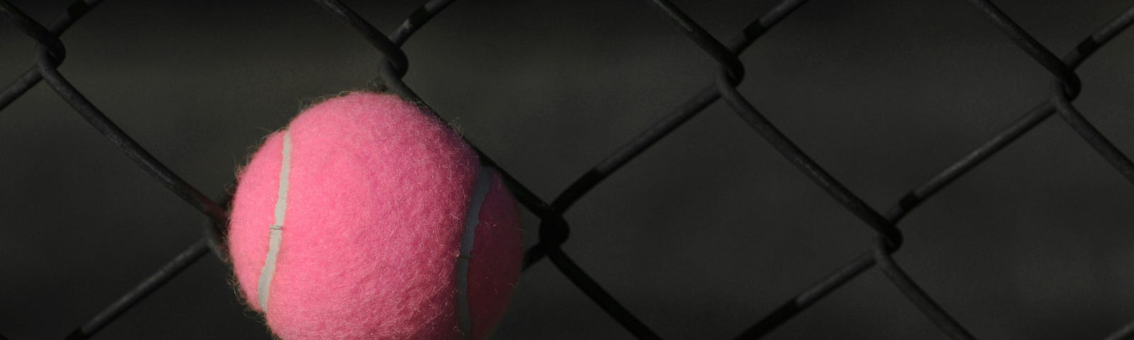 A pink tennis ball stuck on a dark fence.