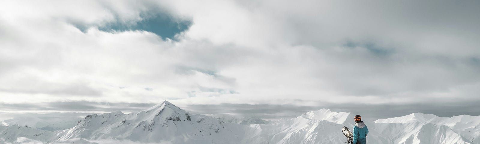 A snowboarder standing on a tall hill looking out at mountain peaks.