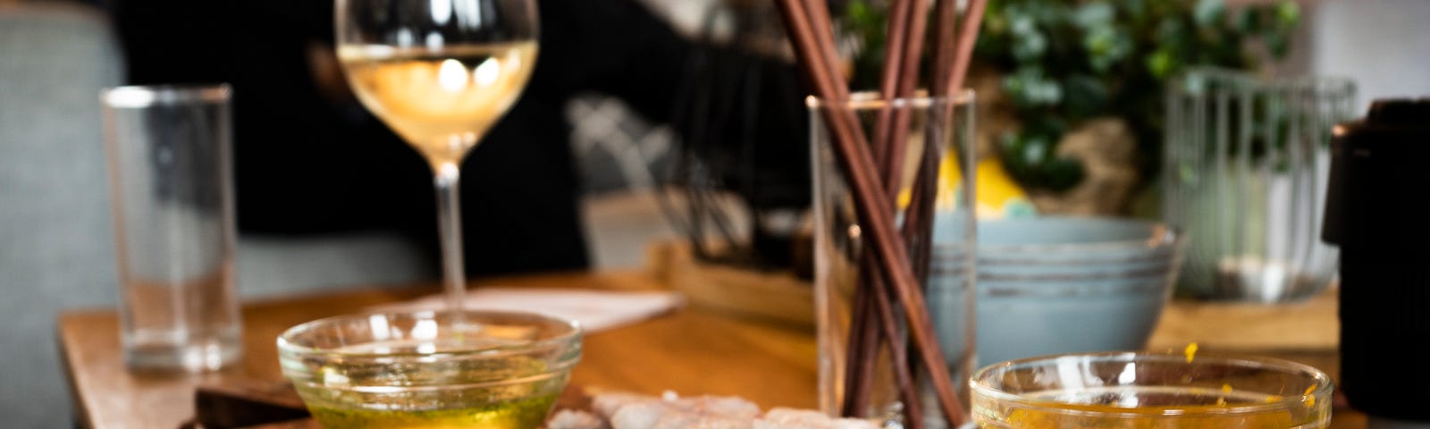 Image shows a wooden plank with pieces of fish it and a glass of wine in the background, with two bowls of fluid to dip the fish in.