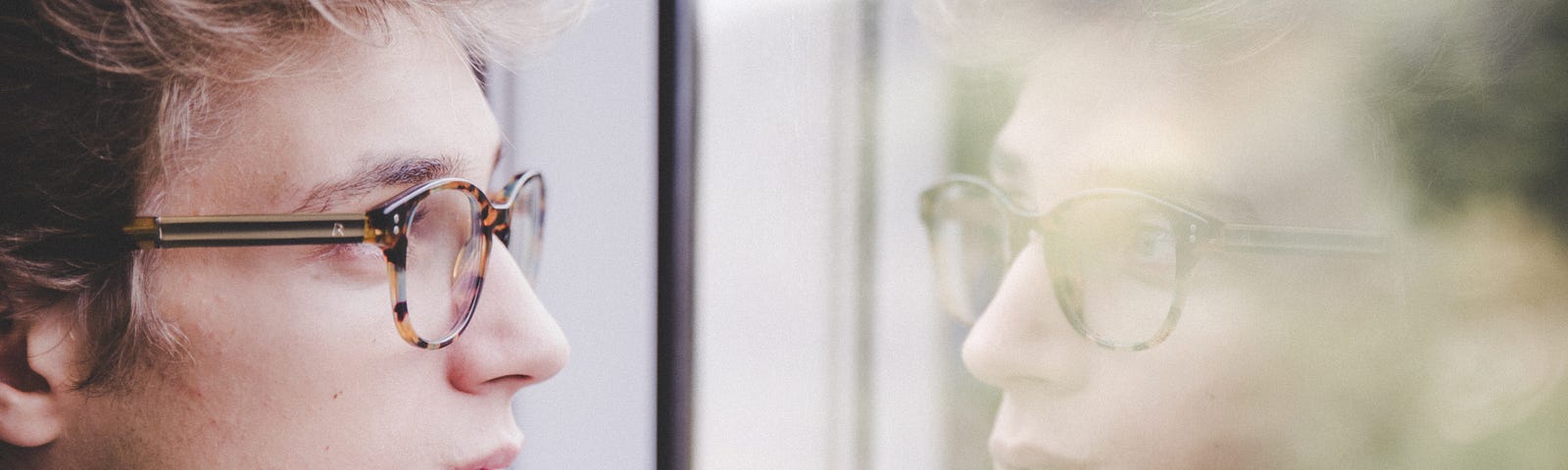 Young man with glasses looking contemplativly at his reflection.