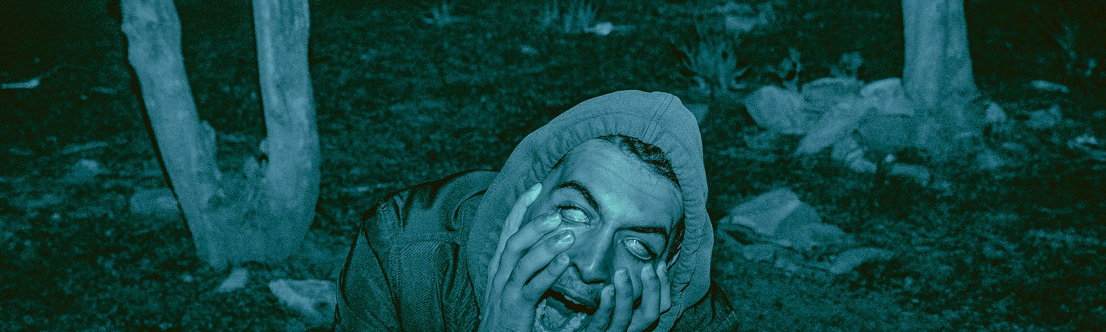 A person with white glazed eyes holding their face and screaming in a dark forest.