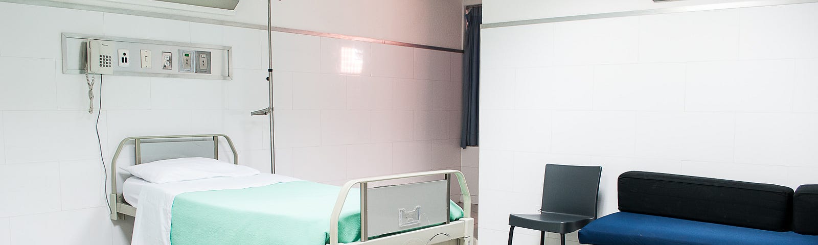 A stark white hospital room with a bed and bench.