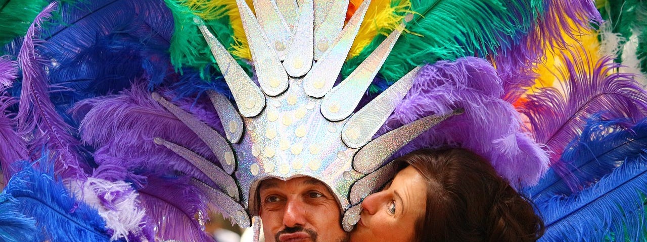 Masculine looking person with a short brown beard/goatee, wearing an iridescent crown and rainbow feather headdress, and a short brown haired femme presenting person kissing them on the cheek, wearing a black tank top