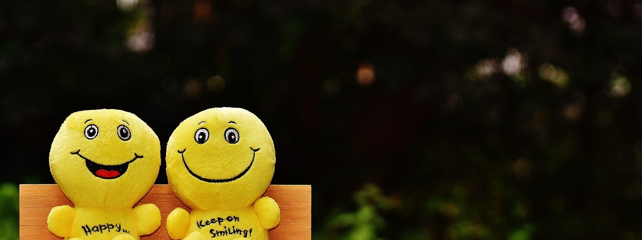 Photo of two yellow smiley faced emoji stuffies, sitting on a wooden bench outside. The one on the left has an open-mouthed smile and says “HAPPY…” on the front, and the other says, “KEEP ON SMILING!”