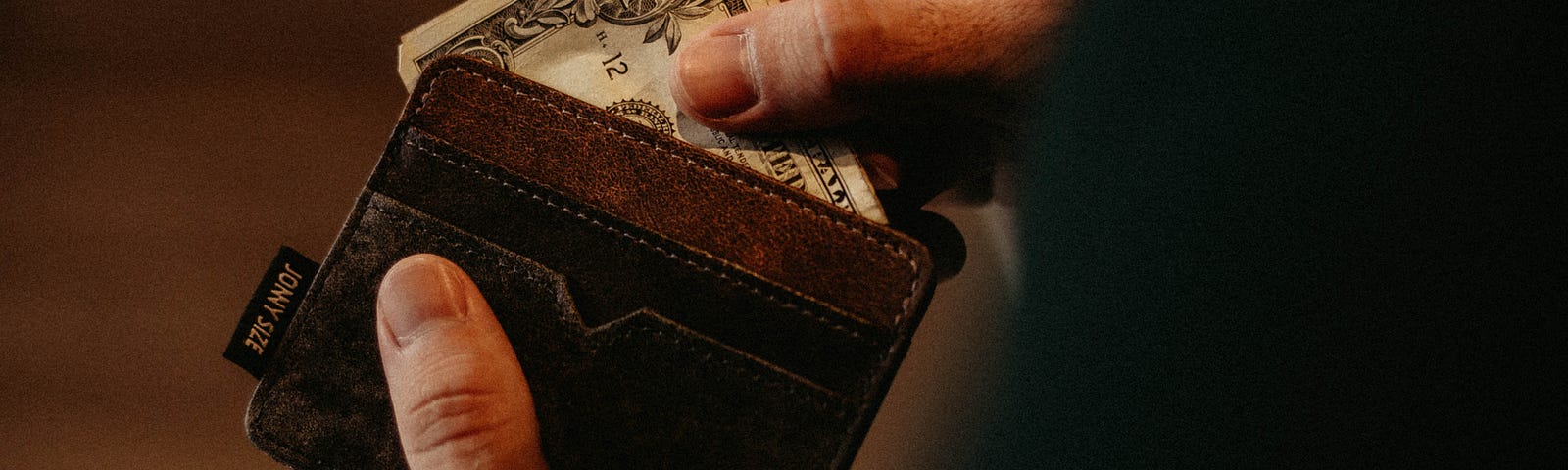 Hands holding a brown leather wallet containing a folded dollar bill