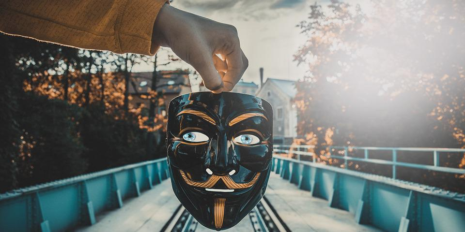 A hand holding a Guy Fawkes mask on a bridge setting.