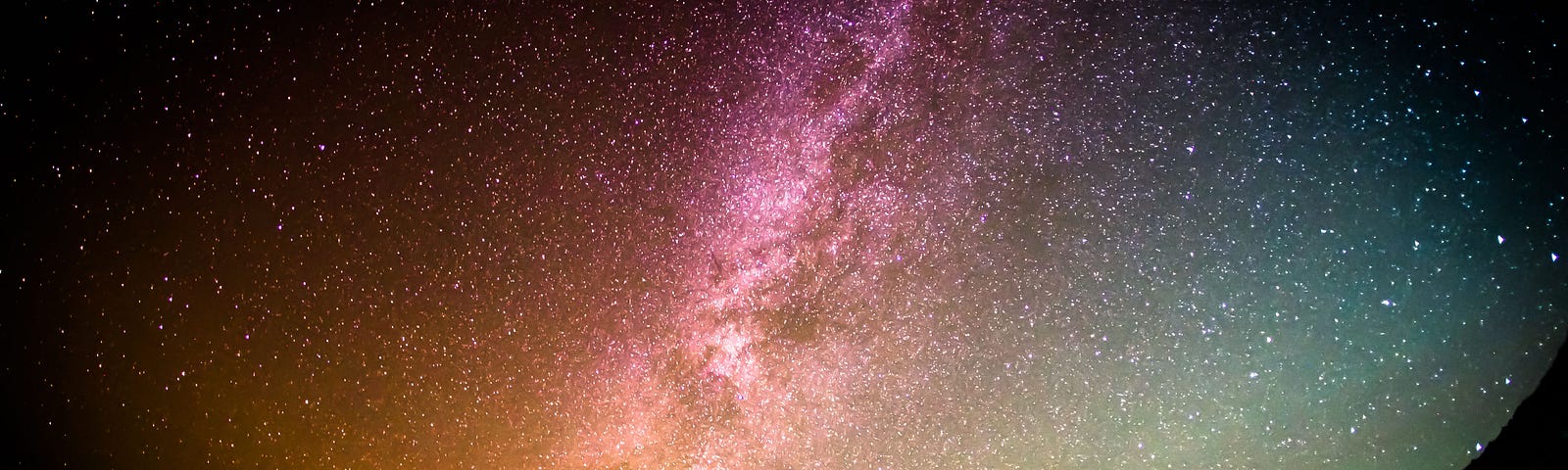 Photo of a rainbow, starry sky against a black background. The sky is visible in round form, taking up most of the image, with the curve towards the bottom of the image. Outside of the curved sky is black. In the centre of the image stands a person, a silhouette with neck bent, looking up towards the cosmos.