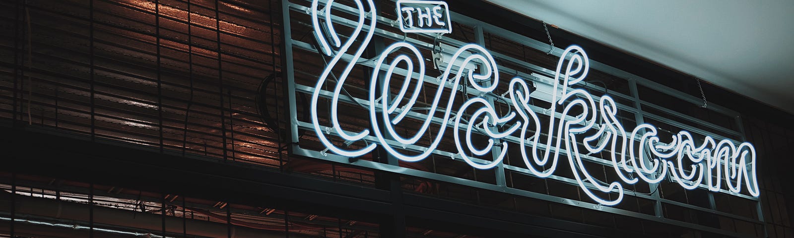 White neon cursive lights that read ‘The workroom’ above the entry to a presumed conference room of sorts.