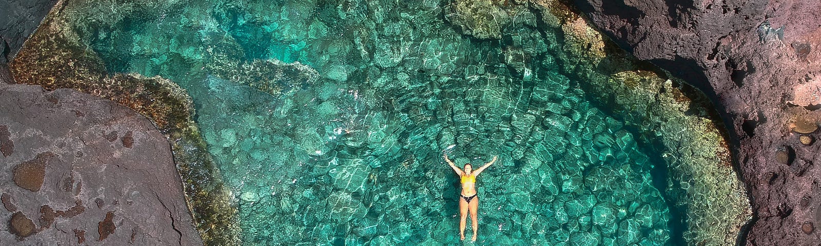 An aerial shot of a woman floating on her back in aquamarine water.