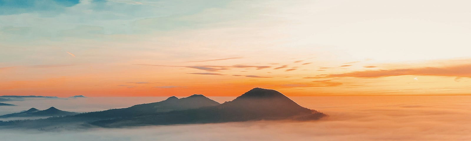 Man standing on precipice eerily overlooking a “sea” of foamy clouds, with distant mountains poking through and highlighted by the rising sun.