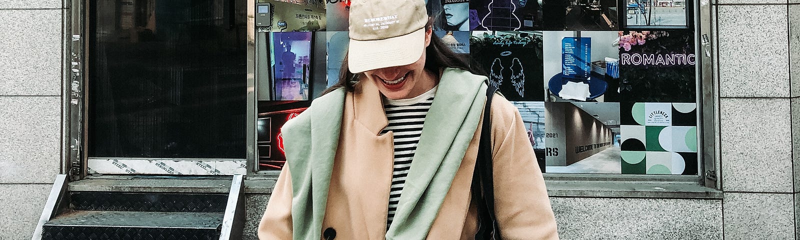 The author wearing a striped shirt, camel coat, with a green hoodie casually walking towards the camera on a street. A camel colored based hat obscures her face so only a smile appears.