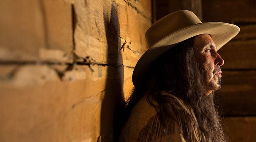 Long-haired man in cowboy hat leaning against log cabin wall