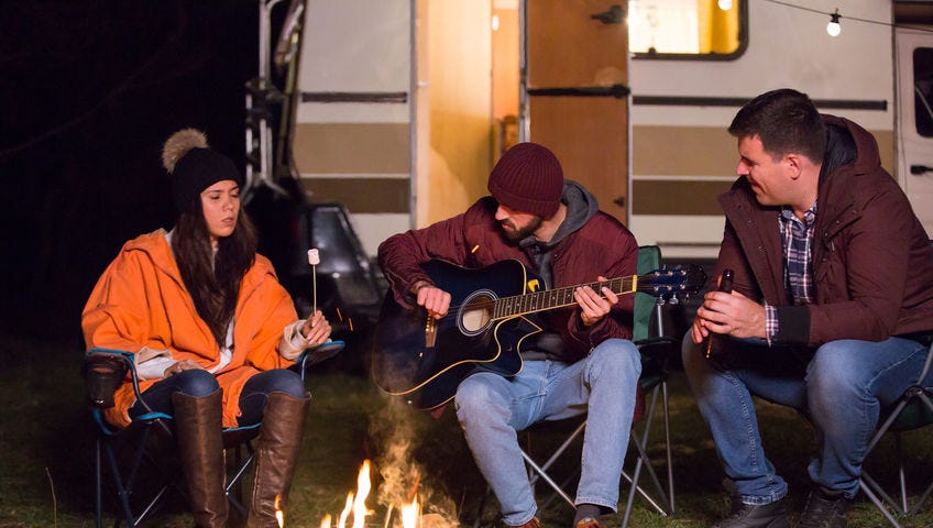 Three people sitting around a campfire in front of an open RV decorated with outdoor lights. One is roasting a marshmallow, one is playing the guitar, and the other is holding a beer.