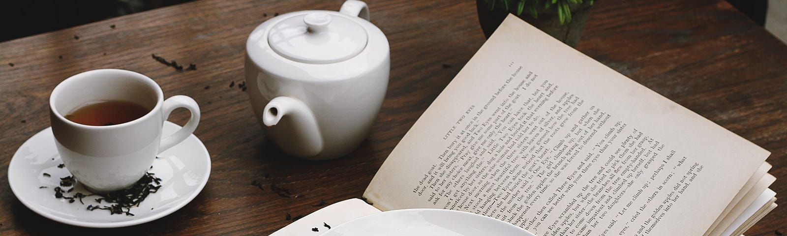 Picture of open book on wooden table with plate of dessert on top and teacup and teapot in the background