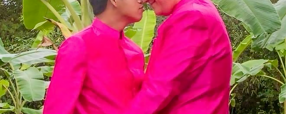 A wedding photo of two men holding each other on a banana plantation, wearing white trousers and bright magenta wedding shirts traditional in Cambodia.