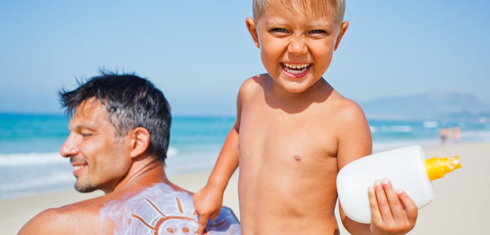Picture of a boy smiling at the camera after he drew a sun in the sunscreen on his father’s back.
