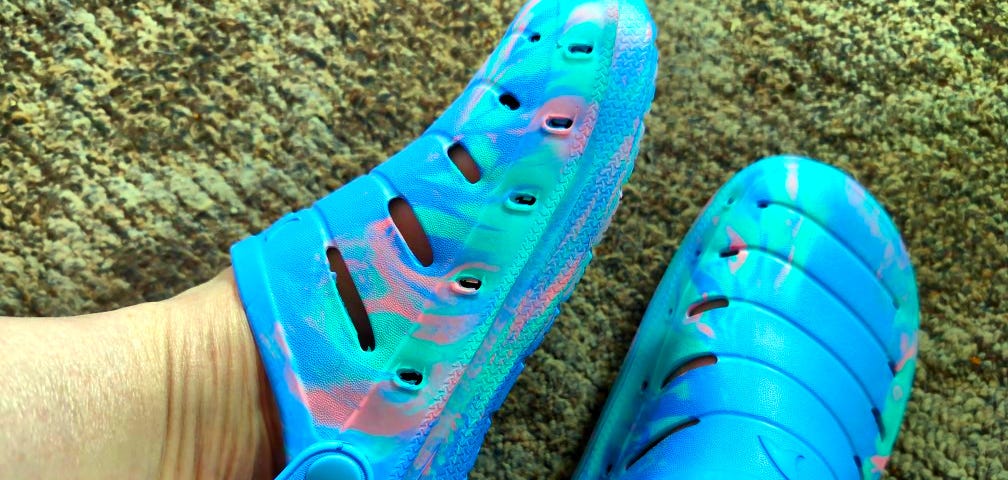 A pair of feet is wearing translucent blue and pink Croc like shoes with multiple cut-outs. The shoes have straps with secure buttons on the sides, and the floor is carpeted.