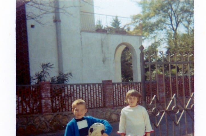 Two children are standing in front of a metal gate, with a building and trees visible in the background. One child is holding a soccer ball and they both appear to be in casual clothing, possibly ready for a game or outdoor activity.