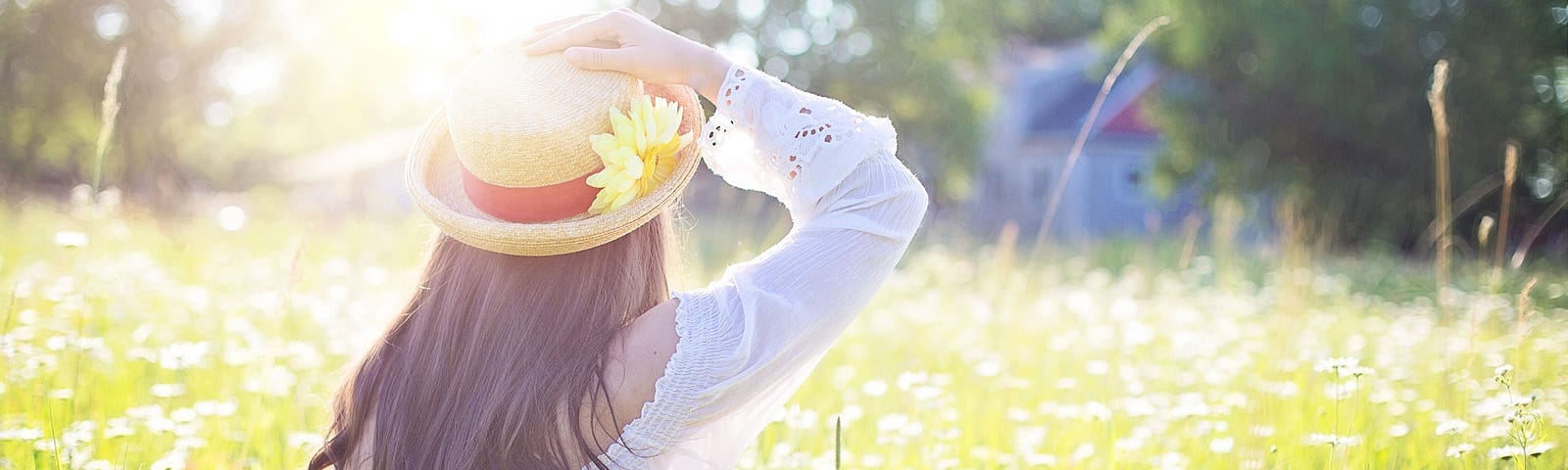 A field of grass and flowers with a person sitting on the ground. The sun shines brightly down on a straw hat with a sunflower on the brim and long brown hair spills out from the hat down the back of a filmy white shirt.