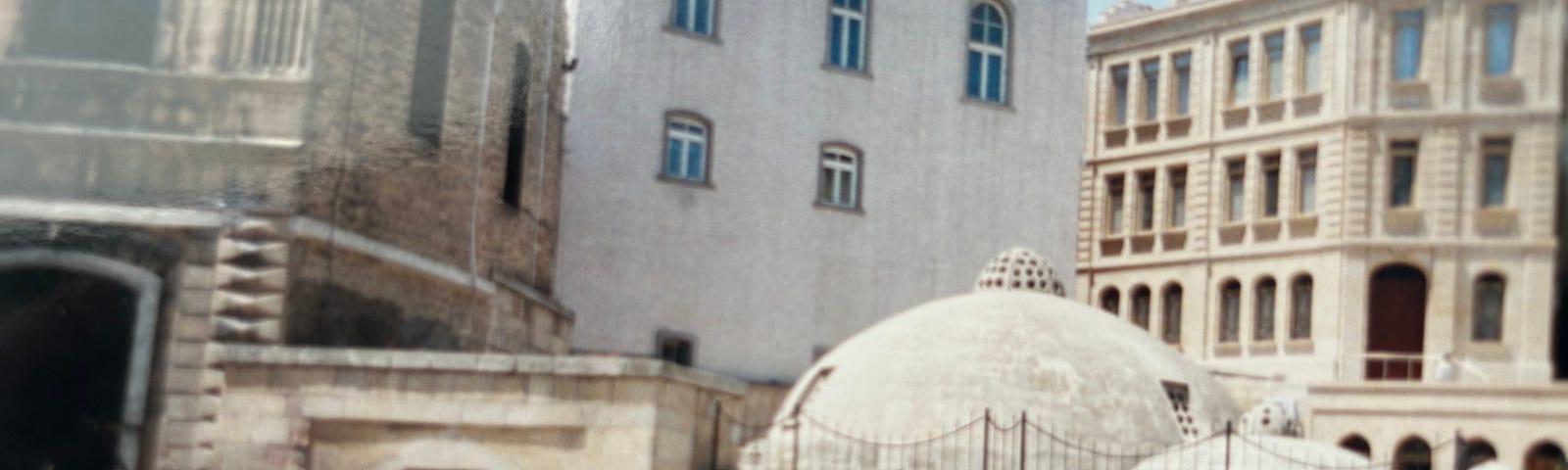 In the foreground is a one-story stone building with domes on top and carpets hanging for sale. In the background are five-story buildings from the 19th century with balconies.