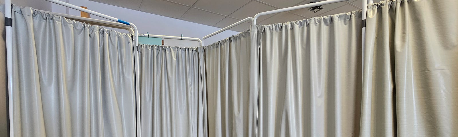 The photo shows a medical room with a divider made of white curtains on a white metal frame. There is a blue medical bed on the left side of the image with a light grey carpet beneath it. The white ceiling has fluorescent lights and square tiles. There are no other people or objects in the room.