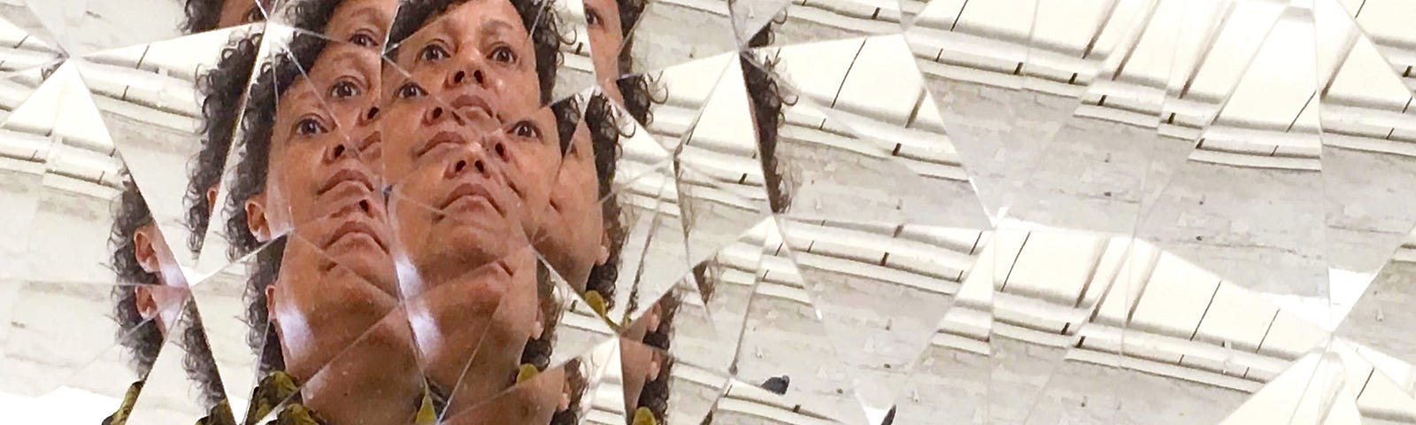 A brown-skin woman gazes at her reflection in a multi-faceted mirror against a white-paneled background. She wears a denim jacket and holds a cellphone camera with a bright floral case. Pieces of her face repeat and ripple out like an aura.