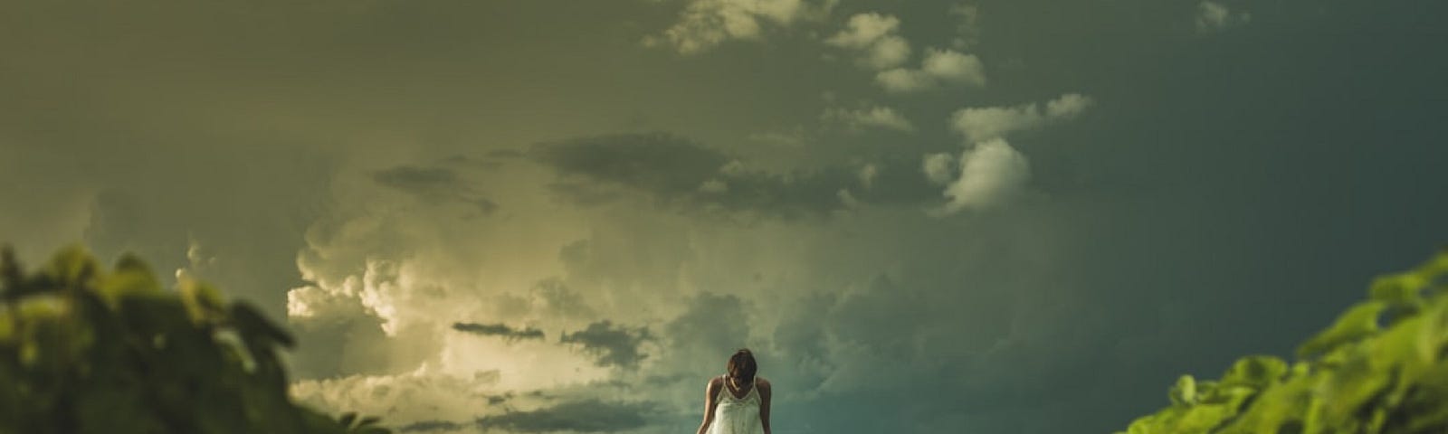 Woman in a white dress, standing in a field surrounded by a stormy sky.