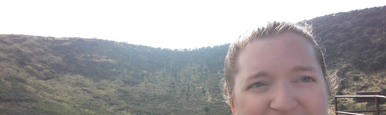 A white woman with brown hair tied back in a ponytail stands at the bottom of a dormant volcano and squints at the camera as she takes a selfie.