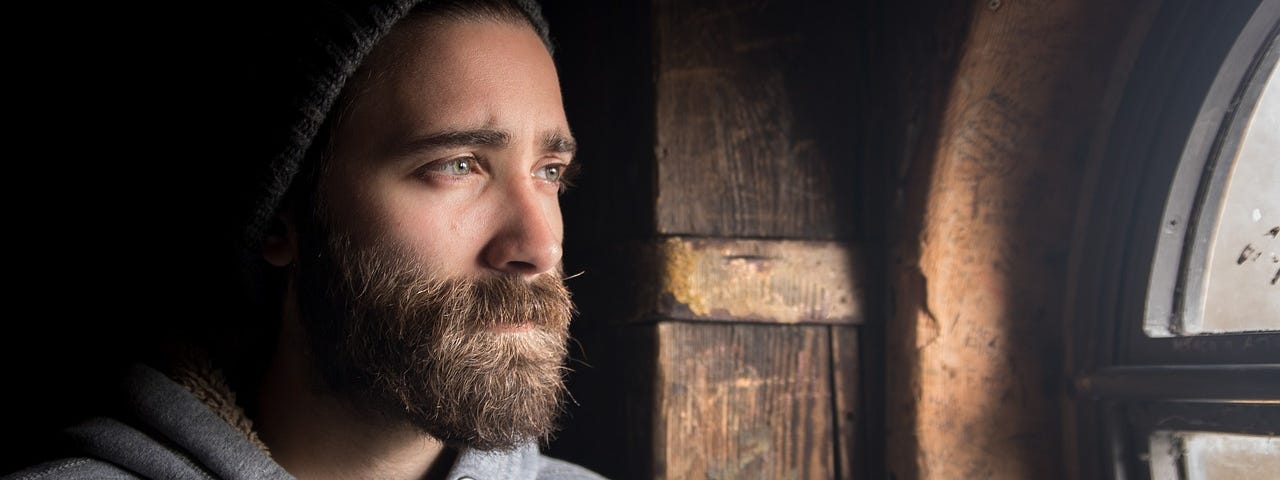 A bearded man wearing a hat looks out the window with a sad expression on his face