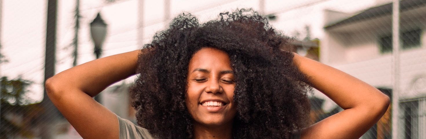 woman smiles with her arms behind her head and eyes closed. She’s outside near trees and buildings.