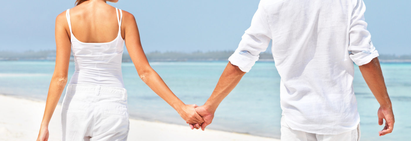 Image of a man and woman walking hand in hand at the ocean. They are looking to one another happy, both dressed in white.