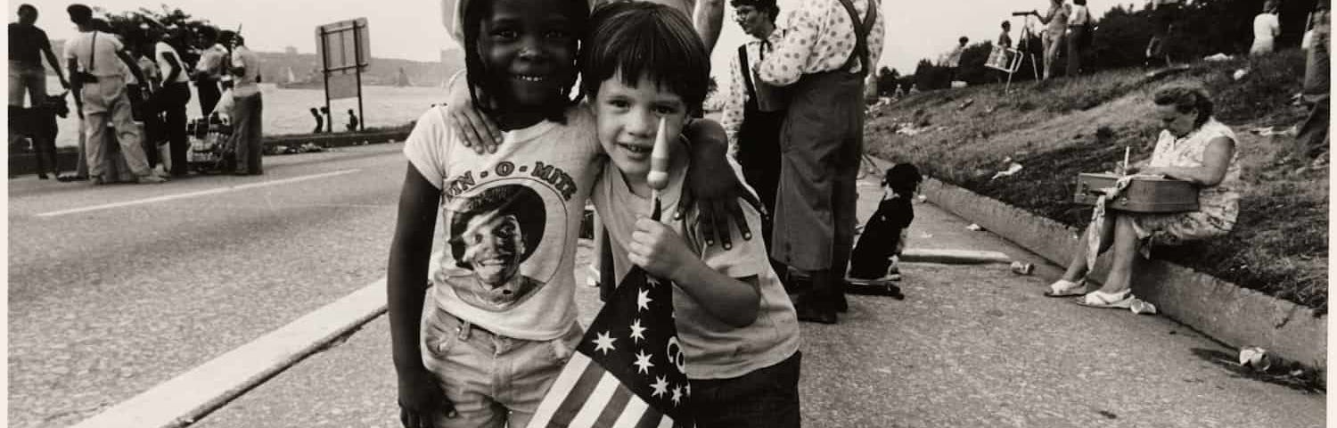 Two boys, about four years old, stand with arms around each other, smiling. One boy is black, one white. One holds an American flag.
