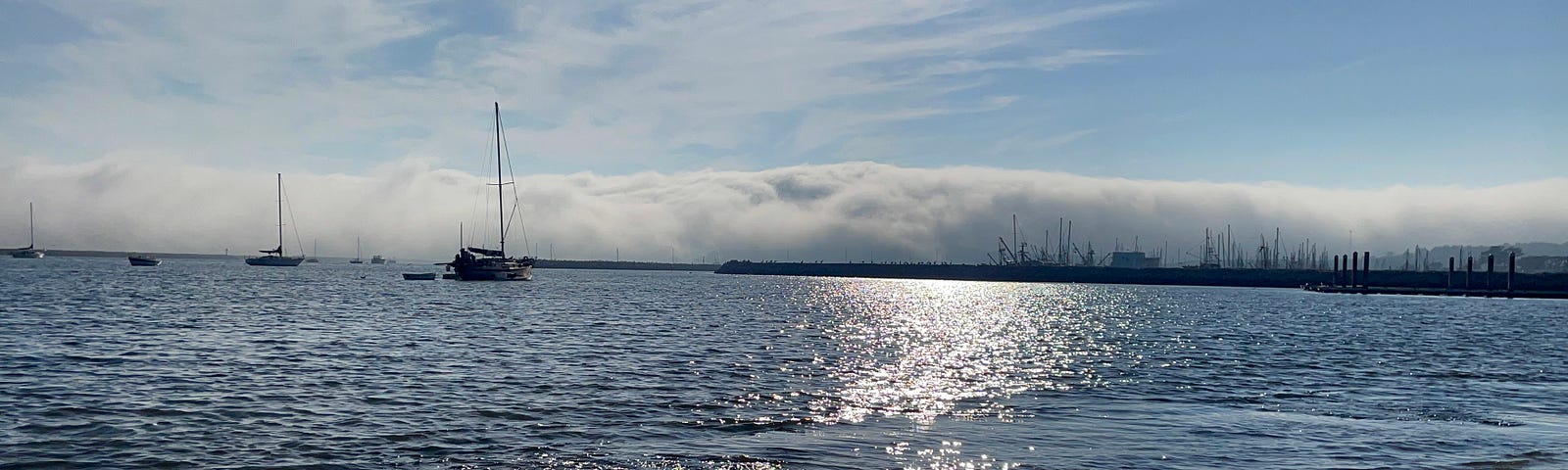 A few boats anchored in Half Moon Bay, as seen from shore, as a large, ominous fog cloud rolls inland from the ocean.