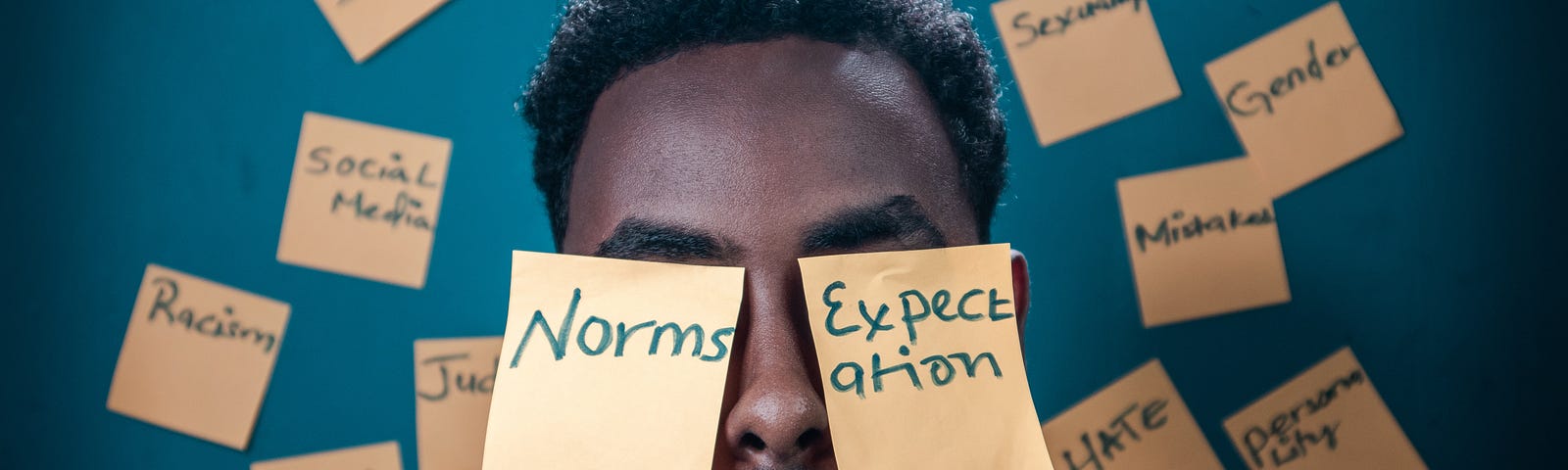 Man with sticky notes on a mans face that say Norms, Expectations, and Society. In the background, there are other sticky notes that say soul, failure, racism, doubt, hate, gender, sexuality, mistaken, personality, justice, and social media