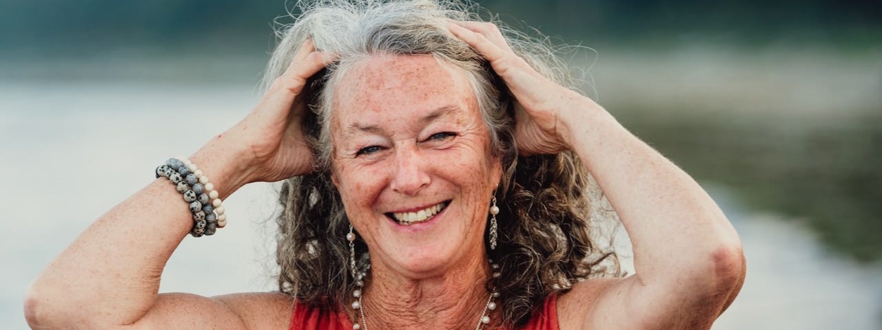 A woman with grey hair has her hands on her head. She is smiling. She is wearing a peach coloured dress and jewelry on wrists and around her neck