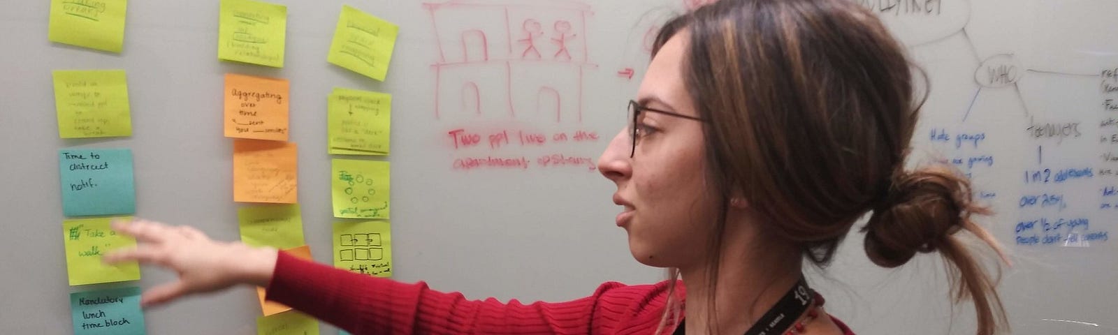 Woman with glasses in a red sweater pointing at Post-its on a whiteboard