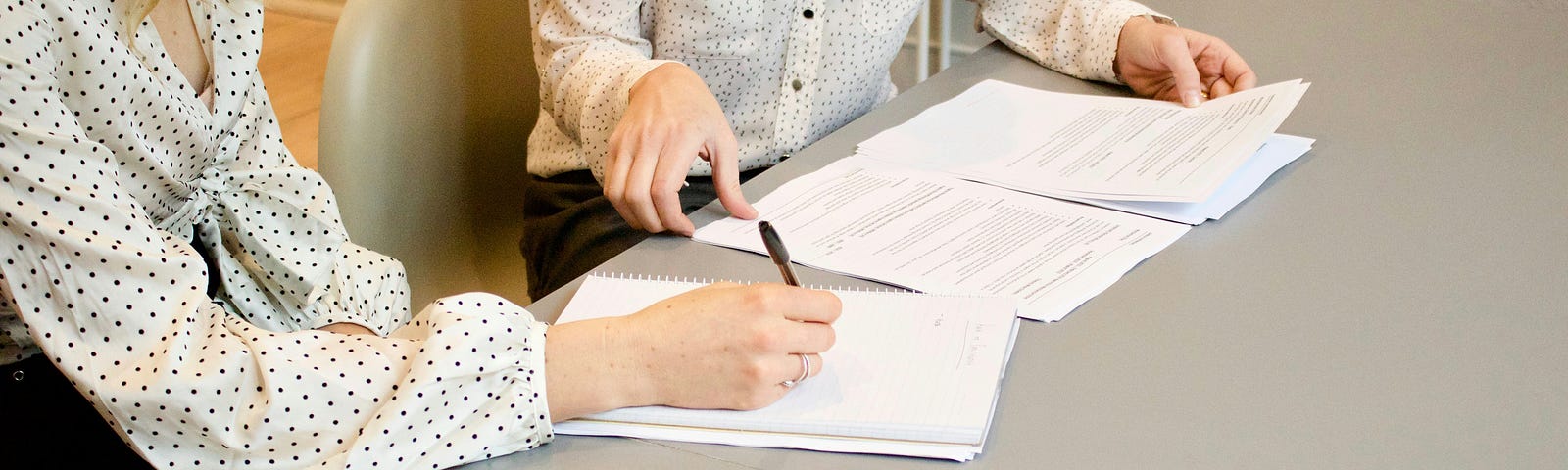 Two women looking over a printed file and taking notes in a business set-up.