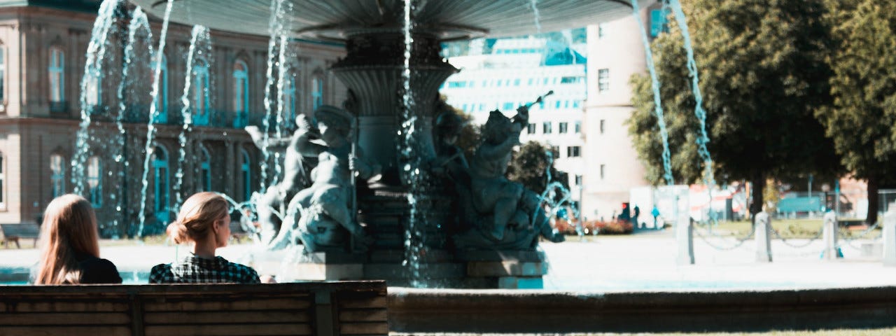 A rear view of two women sitting on a park bench, looking at a large fountain.