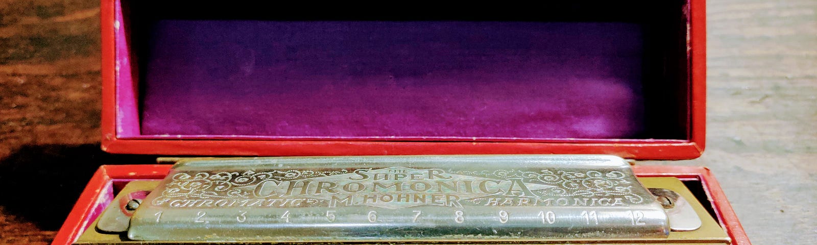 An antique metal Chromonica harmonica sitting in a wooden box with a purple internal lining and red trim.