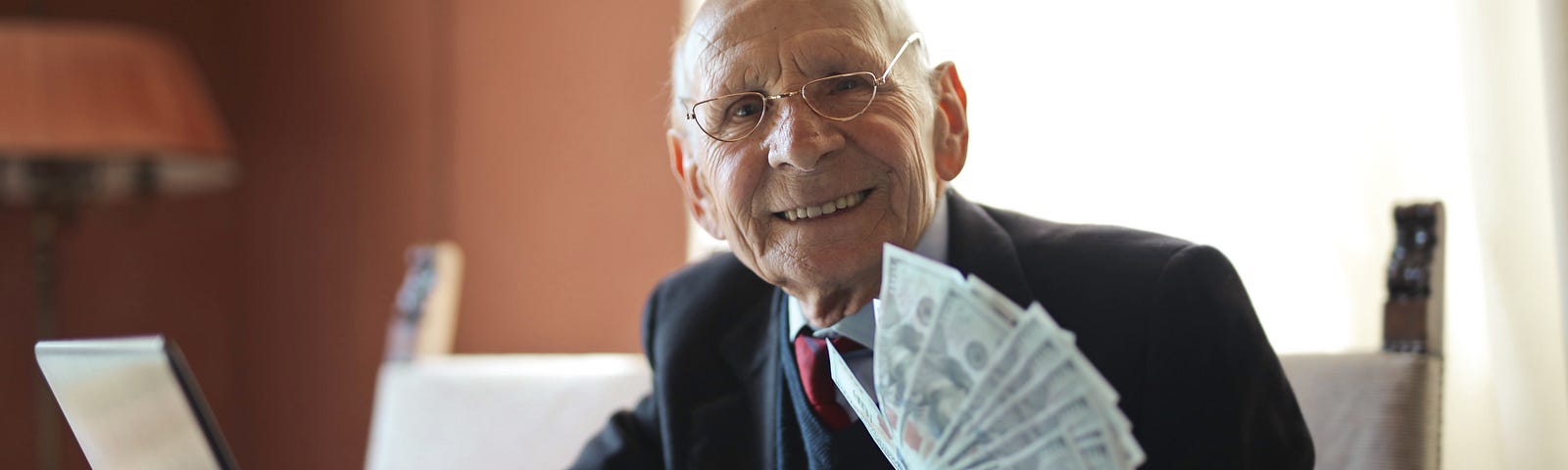happy old man dressed in nice suit holding stack of money 100 dollar bills and sitting next to a computer