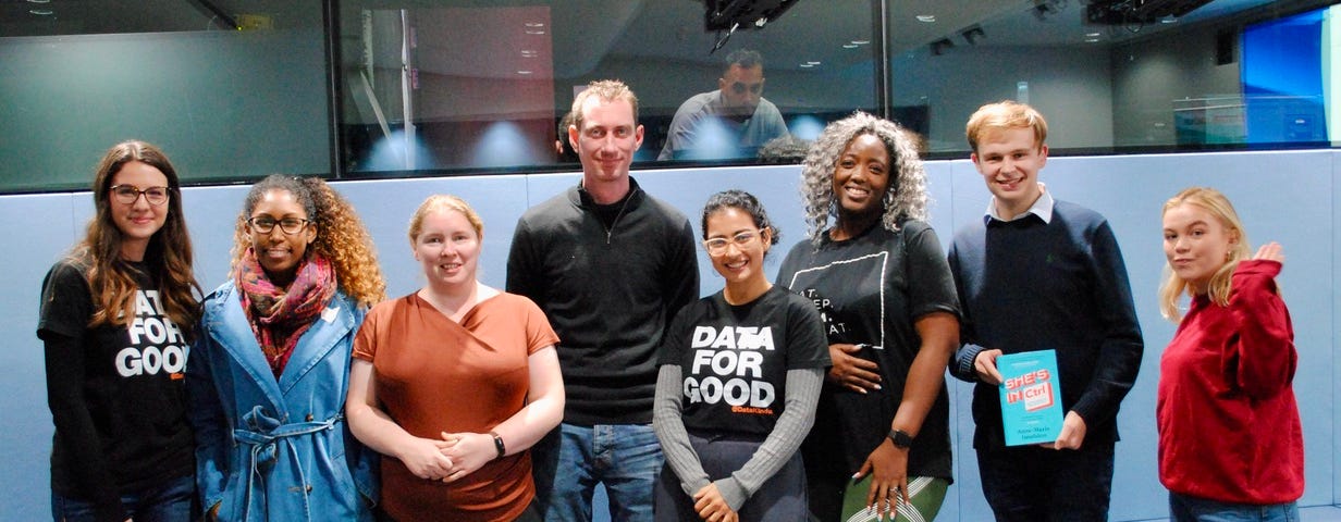 A group of eight adults with broad smiles and relaxed postures, some wearing t-shirts that say ‘Data for Good’, stand together in a conference space