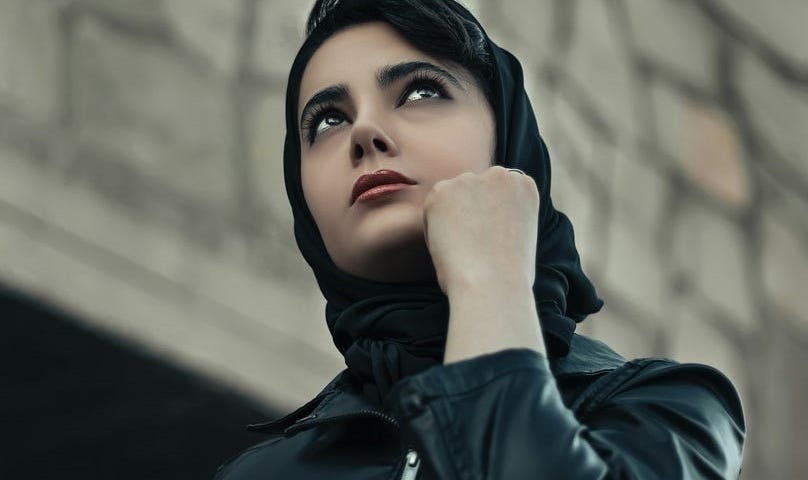 Woman in black leather jacket with black scarf around her head looking thoughtful.