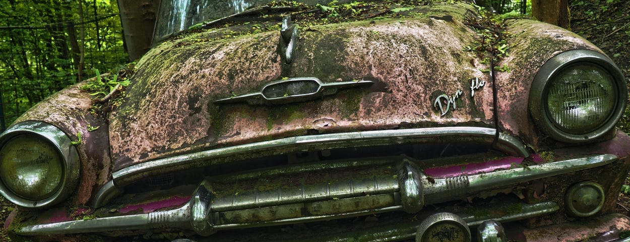 Image: An old car deep in the woods, covered in moss and leaves.