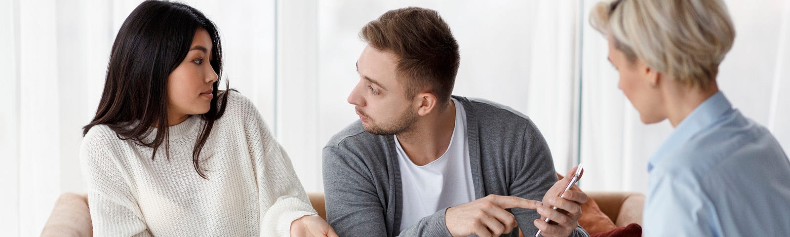 A man and a woman sit together, arguing over something. The man is pointing at a mobile phone, showing her something. A second woman is sat listen to them argue.