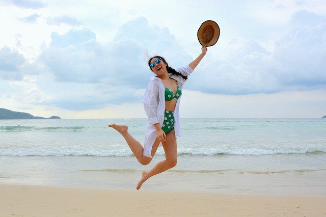 A woman in a two piece bathing suit, holding a hat, and jumping on the beach.