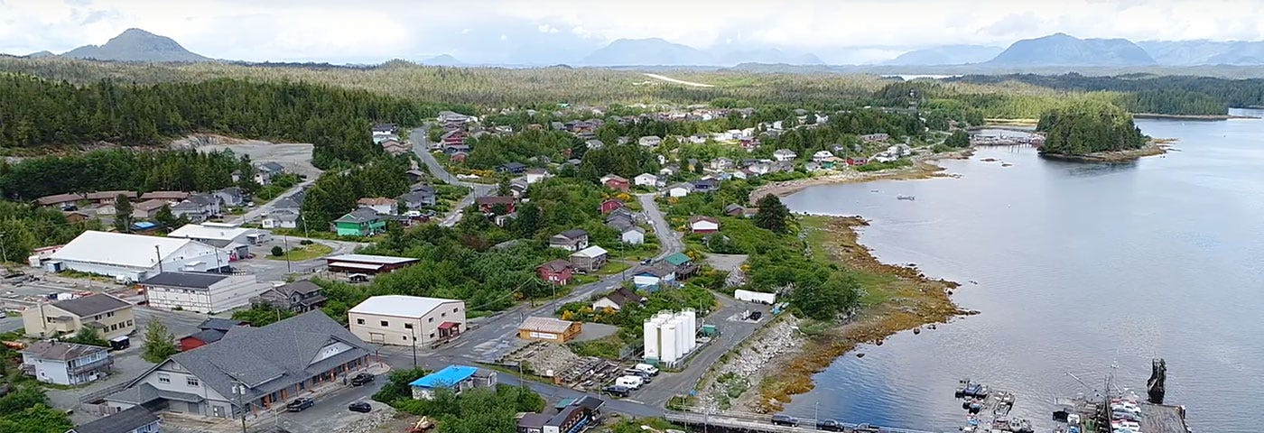 A view of the Haíłzaqv (Heiltsuk) Nation is shown, houses and workplaces surrounded by the green of the trees, streets without traffic, and the great mountains at the end of the photo. The right part of the image shows the sea, and a bridge built over it where some cars travel. Below the bridge, small boats can be seen.