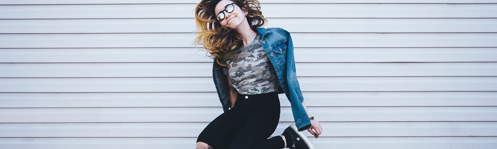 woman wearing black ripped jeans and a denim jacket jumping against a backdrop of a white wall.