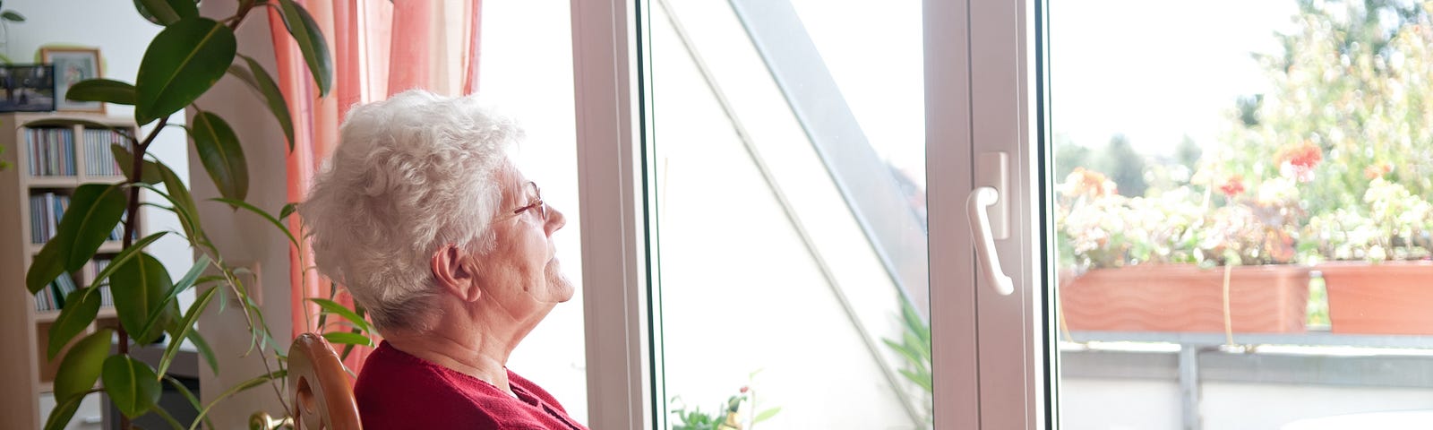 An elderly lady is sitting on a chair in her home and staring out her window.