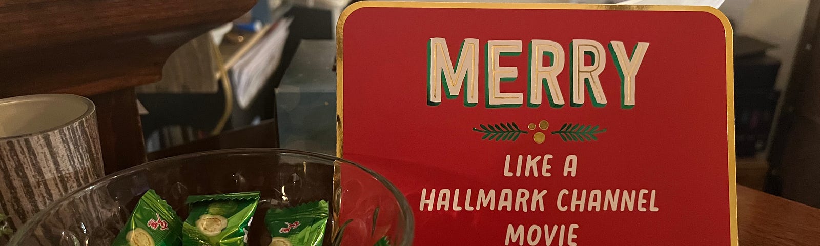 Metal plaque reading “Merry like a Hallmark Channel movie” with a bowl of candy beside it